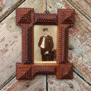 Può includere: Una cornice rettangolare in legno con finitura marrone scuro e un motivo geometrico profondamente intagliato. La cornice contiene una fotografia vintage color seppia di una persona che indossa un abito scuro, un gilet chiaro e un berretto.