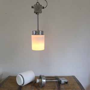 Könnte beinhalten: Vintage Industrie-Pendelleuchte mit zylindrischem, weißem Glasschirm. Die Leuchte hat ein Metallgehäuse und wird von einer an der Decke montierten Anschlussdose abgehängt. Eine zerlegte Leuchte liegt auf einer Holzoberfläche.