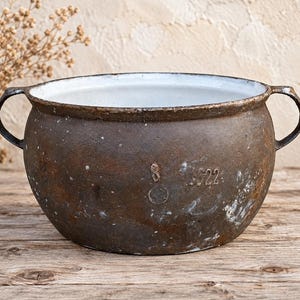 Puede incluir: Una olla redonda y resistente de hierro fundido con dos asas laterales y un interior de esmalte blanco. El exterior tiene un acabado marrón oscuro desgastado con pátina de óxido visible y los números 8 y 1922 en relieve en la parte frontal.