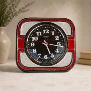 May include: A square Peter Transistor clock with a black face, white numerals, and red second hand. The frame features a textured off-white center with bold red and dark brown borders. The text "peter transistor" and "made in germany" are visible.