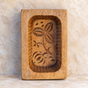May include: A rectangular light brown wooden butter mold featuring a carved floral design with three leaves and a rounded flower base. The interior walls of the mold have vertical fluted grooves, and the wood displays a natural grain texture.