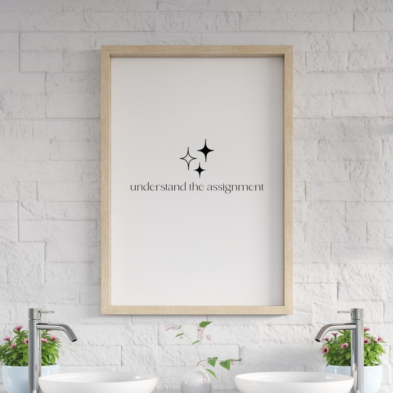 K&ouml;nnte beinhalten: Ein gerahmter Druck mit einem minimalistischen Design mit drei schwarzen Sternen und dem Text "understand the assignment". Der Druck h&auml;ngt an einer wei&szlig;en Backsteinwand.
