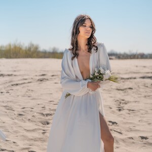 May include: A woman in a white, flowing dress with a plunging neckline stands on a sandy beach. She is holding a bouquet of white flowers.