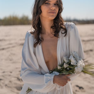 May include: A woman in a white, flowing dress with a plunging neckline holds a bouquet of white flowers. She is standing in a desert landscape.