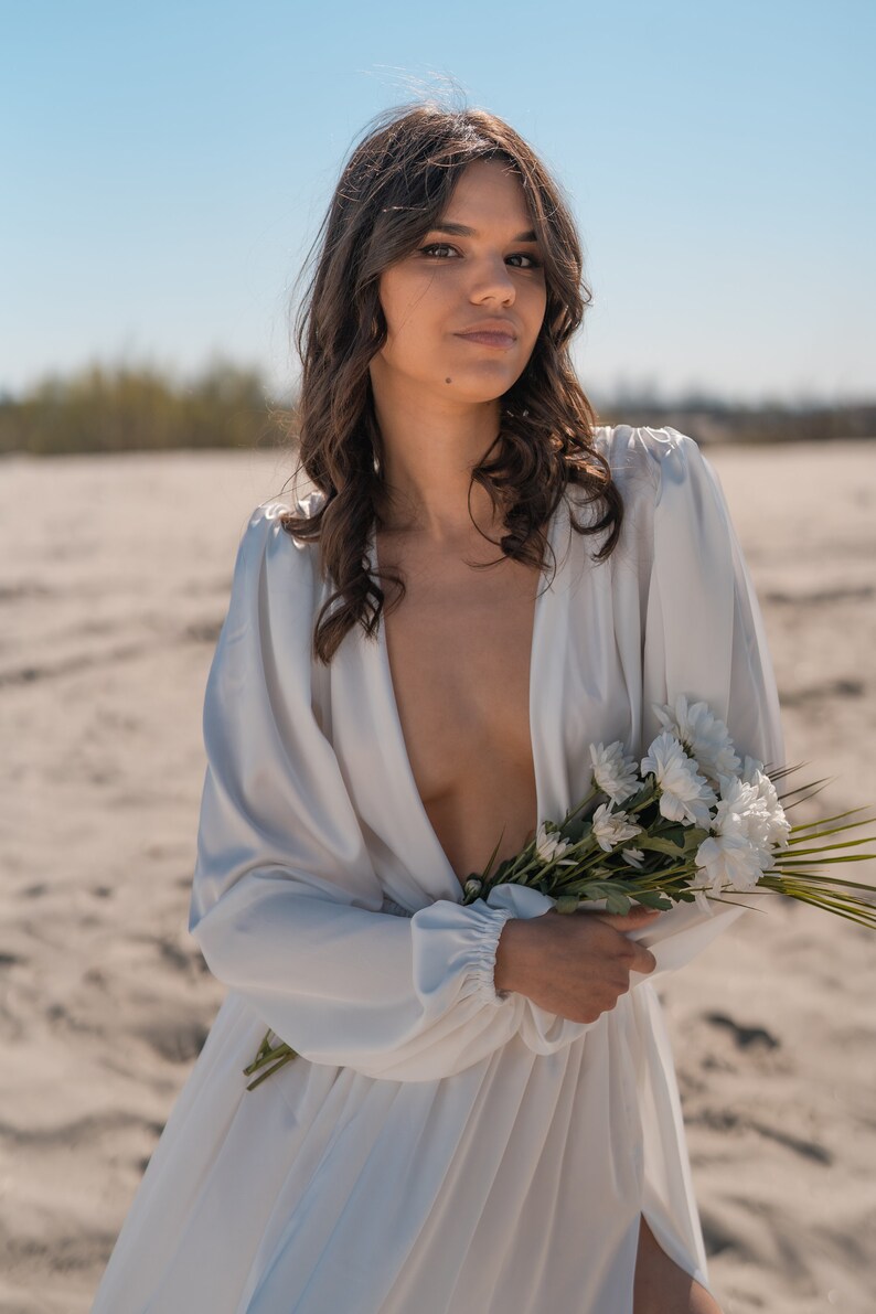 May include: A woman in a white, flowing dress with a plunging neckline holds a bouquet of white flowers. She is standing in a desert landscape.