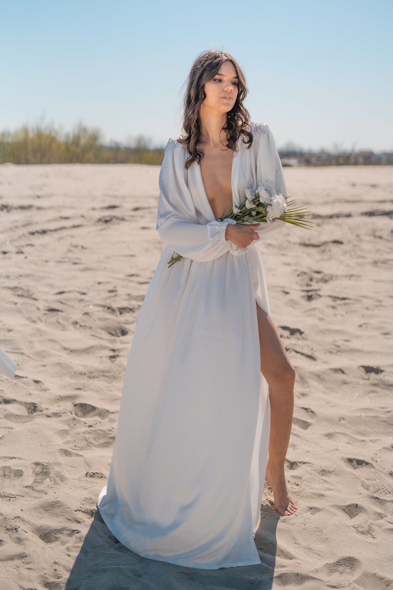 May include: A woman in a white, flowing dress with a plunging neckline stands on a sandy beach. She is holding a bouquet of white flowers.
