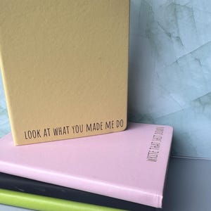 May include: A stack of notebooks in various colors: yellow, pink, black, and green. The yellow notebook on top has the text "LOOK AT WHAT YOU MADE ME DO". The pink notebook has the text "WRITE THAT SHIT DOWN".