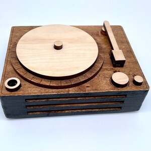 K&ouml;nnte beinhalten: Ein Holz-Plattenspieler mit brauner Oberfl&auml;che. Der Plattenspieler hat einen runden Plattenteller, einen Tonarm und zwei Kn&ouml;pfe. Der Plattenteller ist aus hellem Holz und hat einen dunkelbraunen Rand.