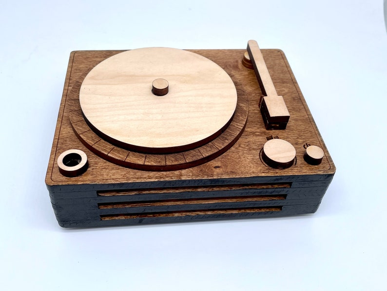 K&ouml;nnte beinhalten: Ein Holz-Plattenspieler mit brauner Oberfl&auml;che. Der Plattenspieler hat einen runden Plattenteller, einen Tonarm und zwei Kn&ouml;pfe. Der Plattenteller ist aus hellem Holz und hat einen dunkelbraunen Rand.