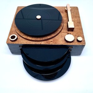K&ouml;nnte beinhalten: Ein Holz-Plattenspieler mit einem schwarzen Plattenteller und einem Stapel von vier schwarzen Vinylplatten. Der Plattenspieler hat einen Holztonarm und einen Holzknopf.