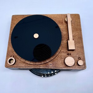 K&ouml;nnte beinhalten: Ein Holz-Plattenspieler mit einer schwarzen Schallplatte, einem Lautst&auml;rkeregler und einem Ein-/Ausschalter. Der Plattenspieler ist aus Holz und hat einen Vintage-Look.