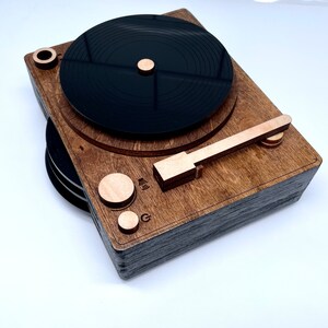 K&ouml;nnte beinhalten: Ein Holz-Plattenspieler mit einer schwarzen Schallplatte, einem Holzarm und einem Holzsockel. Der Plattenspieler hat eine braune Beize und eine schwarze Oberfl&auml;che.