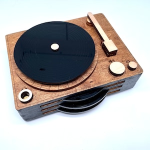 K&ouml;nnte beinhalten: Ein Holz-Plattenspieler mit einer schwarzen Schallplatte und einem Holzarm. Der Plattenspieler hat ein geschichtetes Design mit drei sichtbaren Schichten.