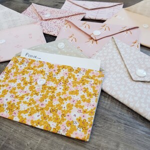 K&ouml;nnte beinhalten: Eine Sammlung von Stoffumschl&auml;gen in verschiedenen Rosa- und Cremet&ouml;nen mit floralen und gemusterten Designs. Jeder Umschlag hat einen wei&szlig;en Druckknopfverschluss. Ein Umschlag ist ge&ouml;ffnet und zeigt einen Zettel mit dem Text "Pssst..."
