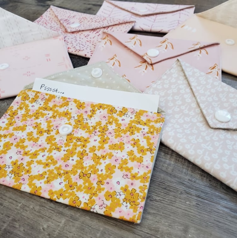 K&ouml;nnte beinhalten: Eine Sammlung von Stoffumschl&auml;gen in verschiedenen Rosa- und Cremet&ouml;nen mit floralen und gemusterten Designs. Jeder Umschlag hat einen wei&szlig;en Druckknopfverschluss. Ein Umschlag ist ge&ouml;ffnet und zeigt einen Zettel mit dem Text "Pssst..."
