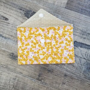 K&ouml;nnte beinhalten: Stofftasche im Umschlagstil mit einem Blumenmuster in Gelb und Rosa auf wei&szlig;em Grund. Die Innenseite ist hellbeige mit einem kleinen Kreuzmuster. Zwei wei&szlig;e Druckkn&ouml;pfe sichern die Klappe.