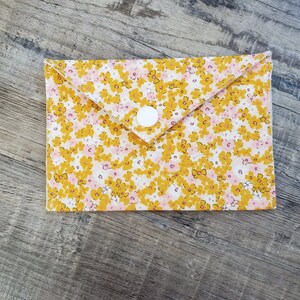 K&ouml;nnte beinhalten: Eine Brieftasche im Umschlagstil mit floralem Muster und wei&szlig;em Druckknopfverschluss. Der Stoff hat ein Muster aus gelben und rosa Blumen auf wei&szlig;em Grund. Die Brieftasche ist rechteckig mit einer gefalteten Klappe.