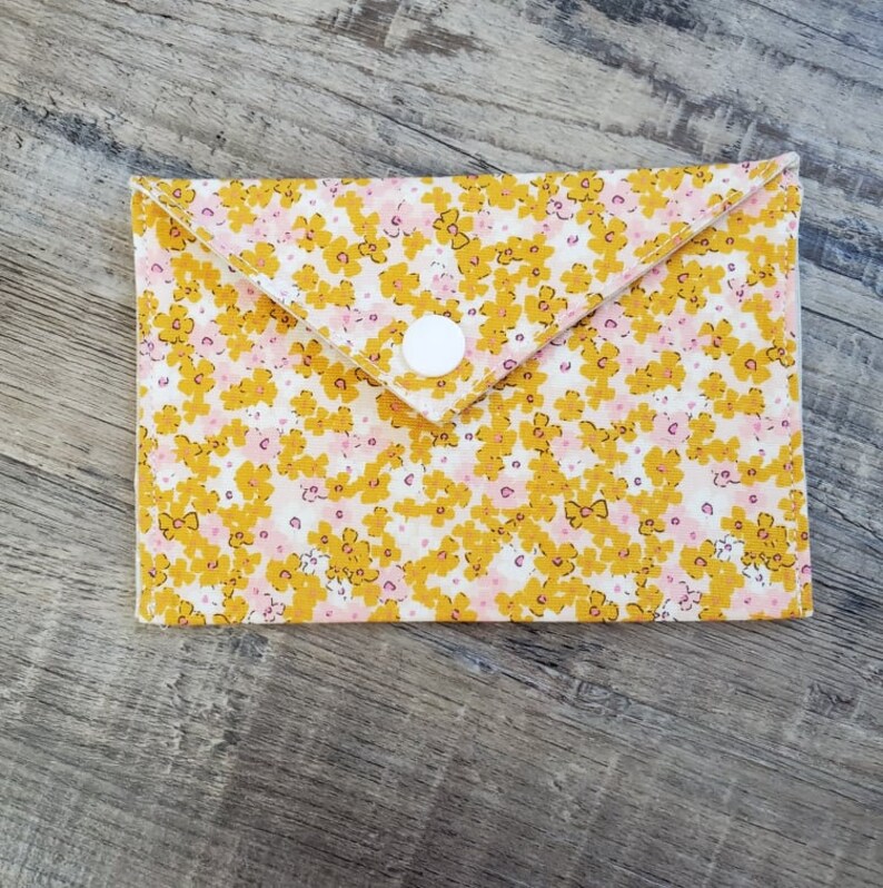 K&ouml;nnte beinhalten: Eine Brieftasche im Umschlagstil mit floralem Muster und wei&szlig;em Druckknopfverschluss. Der Stoff hat ein Muster aus gelben und rosa Blumen auf wei&szlig;em Grund. Die Brieftasche ist rechteckig mit einer gefalteten Klappe.