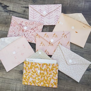 K&ouml;nnte beinhalten: Eine Sammlung von Stoffumschl&auml;gen in Pastellfarben und verschiedenen Mustern, darunter Blumen- und abstrakte Designs. Jeder Umschlag ist mit einem wei&szlig;en Druckknopf verschlossen. Einige Umschl&auml;ge sind ge&ouml;ffnet und zeigen das Innere.