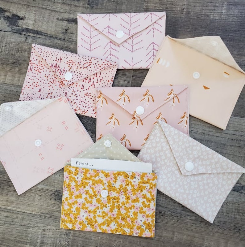 K&ouml;nnte beinhalten: Eine Sammlung von Stoffumschl&auml;gen in Pastellfarben und verschiedenen Mustern, darunter Blumen- und abstrakte Designs. Jeder Umschlag ist mit einem wei&szlig;en Druckknopf verschlossen. Einige Umschl&auml;ge sind ge&ouml;ffnet und zeigen das Innere.