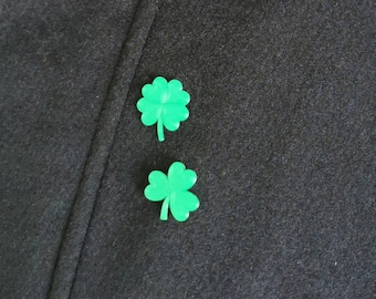 May include: Two bright green shamrock pins are displayed on a dark navy blue fabric. The shamrocks are a vibrant green color and are positioned near each other. The fabric has a textured appearance.