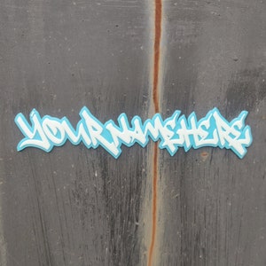 May include: A blue and white graffiti-style sticker with the text "YOUR NAME HERE" on a gray textured surface.
