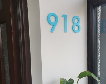 May include: A close-up of a house number sign with the number 918 in bright blue on a white wall.