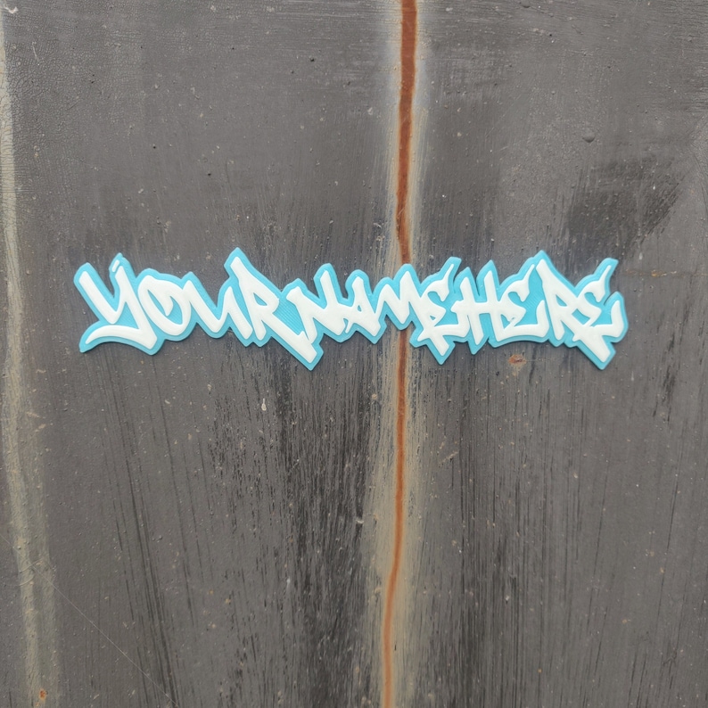 May include: A blue and white graffiti-style sticker with the text "YOUR NAME HERE" on a gray textured surface.