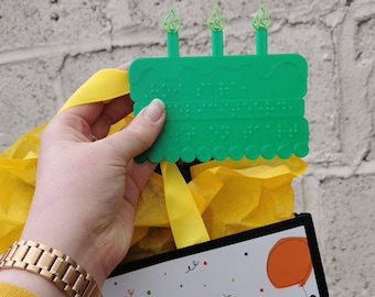 May include: A green birthday cake with three candles, made for a visually impaired person, with braille lettering on the cake. The cake is attached to a yellow ribbon.