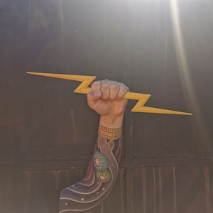 May include: A person's arm with a colorful tattoo is holding a wooden lightning bolt prop. The arm is bent at the elbow and the hand is gripping the prop.