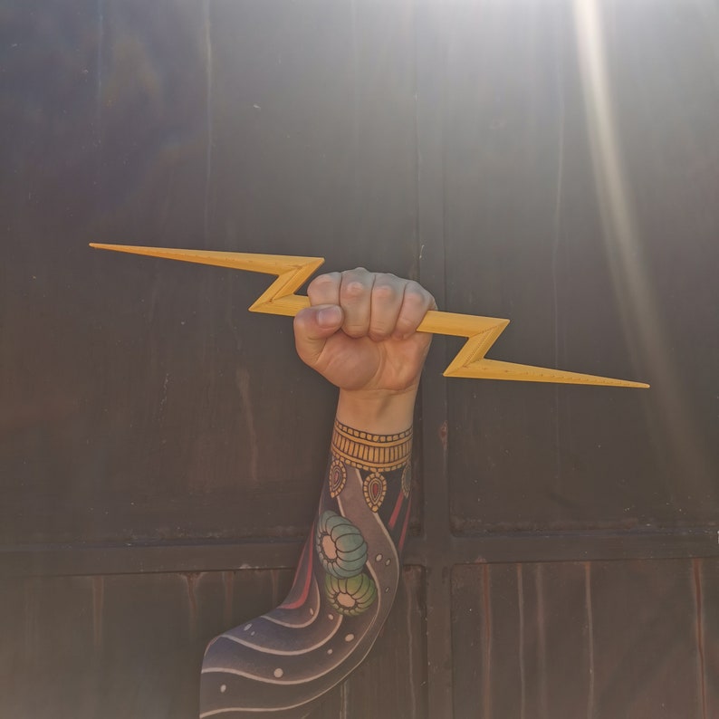May include: A person's arm with a colorful tattoo is holding a wooden lightning bolt prop. The arm is bent at the elbow and the hand is gripping the prop.