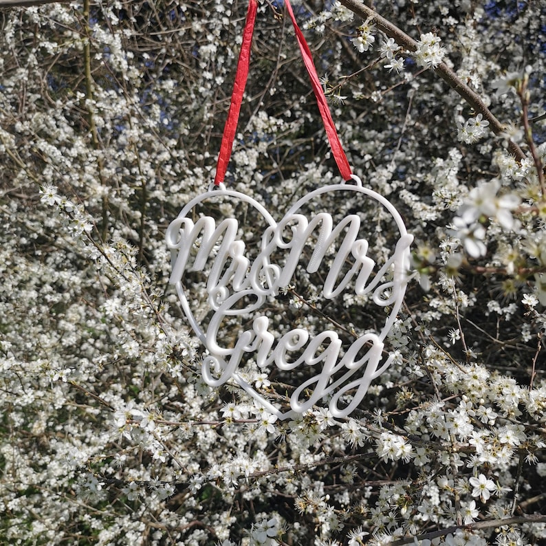 May include: White heart-shaped sign with the text "Mr & Mrs Gregg" hanging from a red ribbon against a background of white flowers.