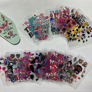 May include: Assortment of clear plastic sheets with colorful, printed designs. The designs include butterflies, hearts, stars, and text such as "Book Lover", "Magic", and "It's Okay to be Off", among others. There is also a green keychain with a butterfly design and the text "Magic Mom Butterfly".