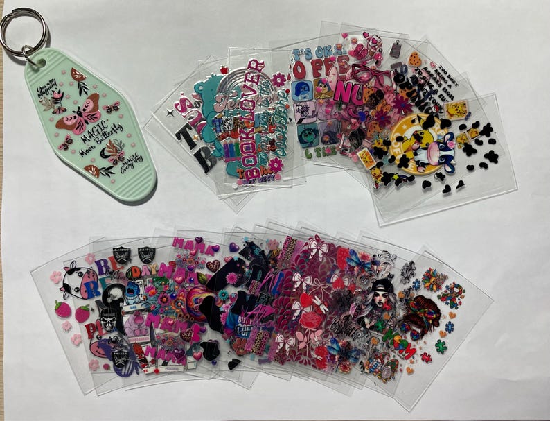 May include: Assortment of clear plastic sheets with colorful, printed designs. The designs include butterflies, hearts, stars, and text such as "Book Lover", "Magic", and "It's Okay to be Off", among others. There is also a green keychain with a butterfly design and the text "Magic Mom Butterfly".