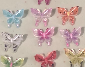 May include: A collection of ten colorful butterfly-shaped embellishments. The butterflies are in various colors, including pink, blue, orange, and green. Each butterfly has a translucent, glittery appearance, with detailed wing patterns.