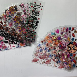 May include: A set of clear plastic sheets with colorful stickers and designs. The stickers feature flowers, hearts, butterflies, and other whimsical designs. One sheet has the text "Livin that Teacher Life" and "Coffee Teacher Powers".