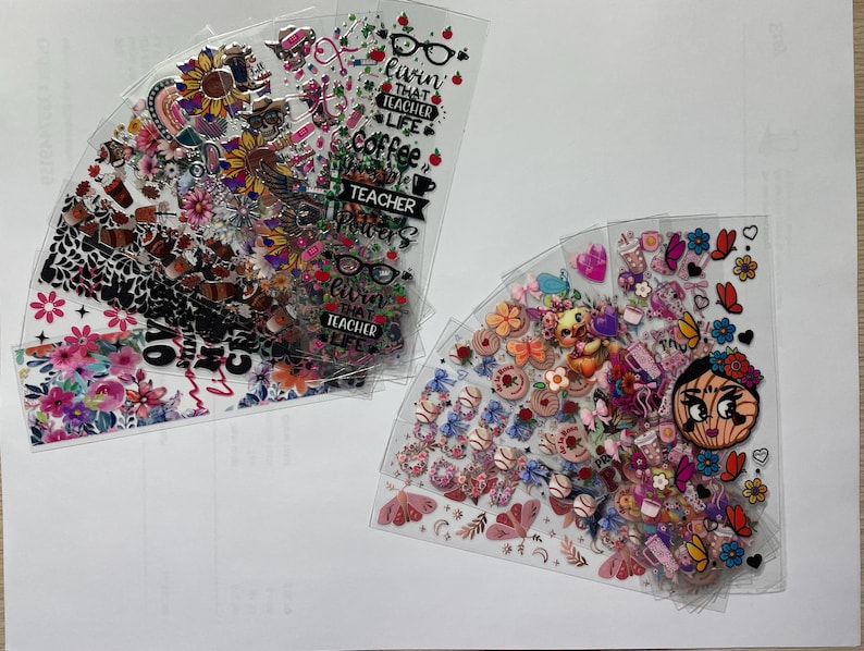 May include: A set of clear plastic sheets with colorful stickers and designs. The stickers feature flowers, hearts, butterflies, and other whimsical designs. One sheet has the text "Livin that Teacher Life" and "Coffee Teacher Powers".
