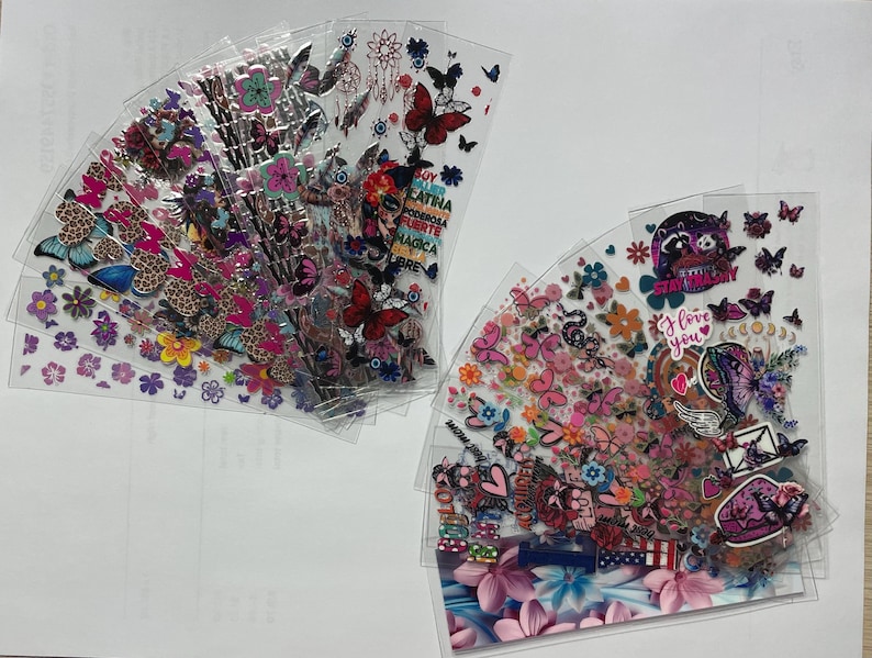 May include: A set of 10 clear plastic sheets with colorful butterfly, flower, and heart stickers. The stickers are arranged in a fan shape and feature various designs, including butterflies, flowers, hearts, and the words "Stay Strong" and "I Love You".