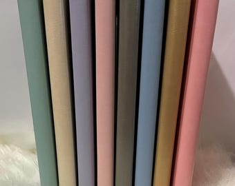 May include: A collection of eight pastel-colored journals, standing upright. The colors include shades of green, beige, lavender, pink, gray, blue, gold, and a brighter pink. The journals are arranged in a row, with their spines facing forward.