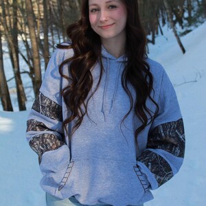 May include: A light gray hoodie with a front pocket and a drawstring hood. The sleeves feature a camouflage pattern. The person is wearing blue jeans. The background is a snowy forest.