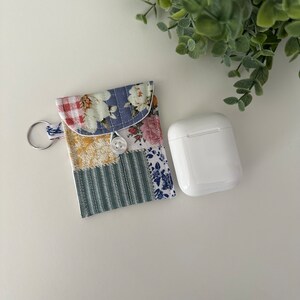 May include: A patchwork fabric case with a zipper closure and a key ring. The case is designed to hold a small electronic device, such as a pair of wireless earbuds. The case is sitting on a white surface next to a white wireless earbud case.