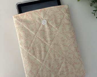May include: Cream quilted tablet sleeve with a pink floral design. The sleeve features a button closure and a diamond stitch pattern. A tablet is partially visible inside the sleeve.