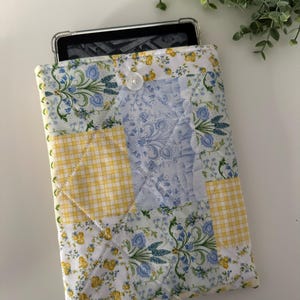 May include: A quilted tablet case with a patchwork design featuring yellow and blue floral patterns. The case has a button closure.