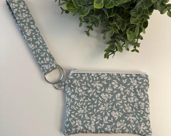 Op de afbeelding: Een kleine, gewatteerde portemonnee met een polsbandje. De portemonnee heeft een gedempte teal kleur met een wit bloemenpatroon. Het heeft een zilveren rits en een zilveren metalen ring bevestigd aan de riem.