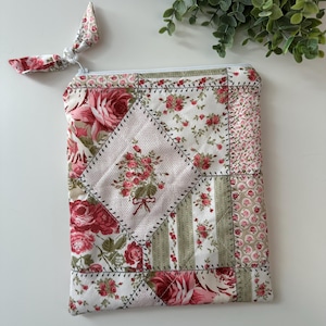 May include: A quilted floral pouch with a zipper closure and a matching fabric pull. The patchwork design features red roses, green stripes, and a central diamond with a floral bouquet. The pouch is approximately 8 inches wide and 10 inches tall.