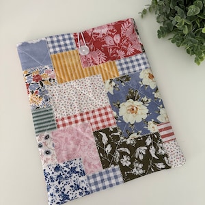 May include: A patchwork tablet sleeve featuring a variety of floral, gingham, and striped fabric squares in shades of blue, red, yellow, and green. The sleeve is secured with a white button and elastic loop. The quilted design adds texture and visual interest.