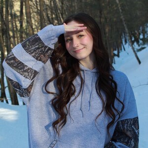 May include: A gray hoodie with camouflage patterned sleeves. The hoodie has a front pocket and a drawstring hood. The sleeves feature a woodland camouflage pattern. The person in the image is wearing the hoodie outdoors in a snowy environment.