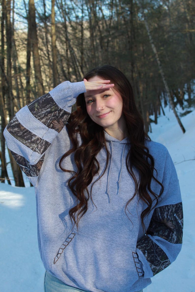 May include: A gray hoodie with camouflage patterned sleeves. The hoodie has a front pocket and a drawstring hood. The sleeves feature a woodland camouflage pattern. The person in the image is wearing the hoodie outdoors in a snowy environment.
