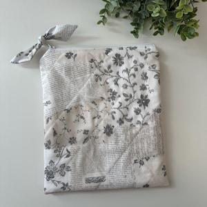 May include: A white quilted pouch with a zipper closure and a fabric bow. The pouch features a floral and newspaper print in gray and white. The design includes a variety of floral patterns and text from old newspapers.
