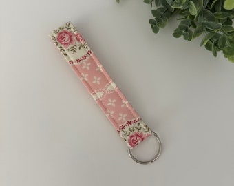 May include: A pink fabric keychain wristlet with a floral and geometric pattern. The wristlet features a silver metal key ring and is decorated with roses and small white flowers. The design includes a mix of pink, white, and red colors.